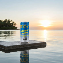 Mojo coconut water and pineapple juice can by the ocean at sunset, tropical beach background
