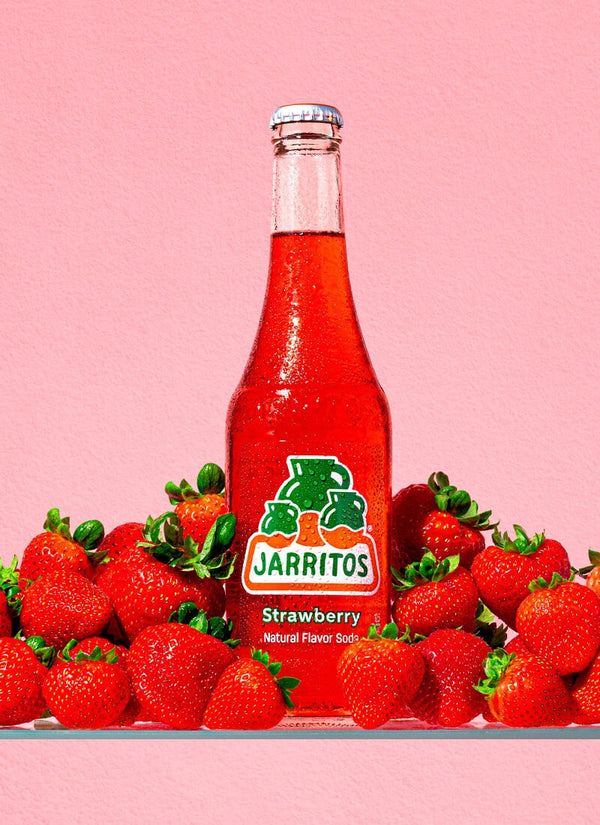 A bottle of Jarritos Strawberry natural flavor soda surrounded by fresh strawberries on a solid pink background.