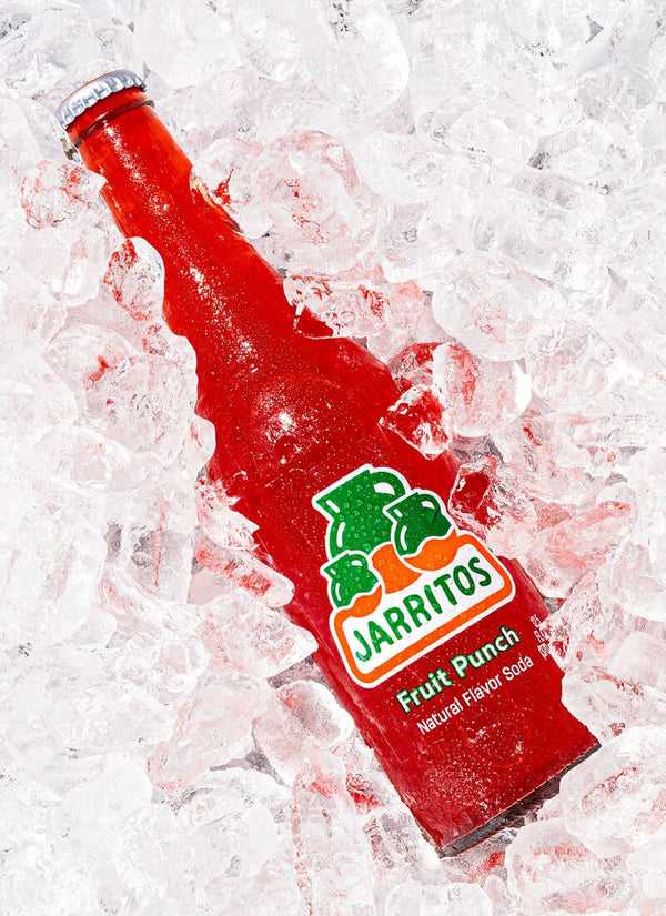 A red Jarritos Fruit Punch soda bottle lying diagonally on a bed of crushed ice. The label features the green and orange Jarritos logo with 'Fruit Punch' and 'Natural Flavored Soda' printed below.
