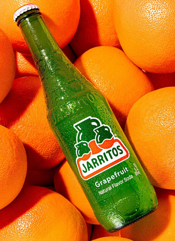 A green glass bottle of Jarritos Grapefruit soda surrounded by fresh orange fruits, with the brand logo clearly visible on the front label.