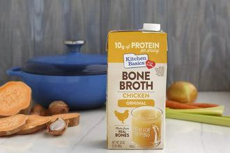 Chicken bone broth carton on kitchen counter with sweet potato, onion, celery, and blue pot