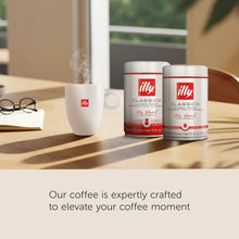 Two illy Classico coffee cans and a steaming coffee mug on a wooden table with sunlight