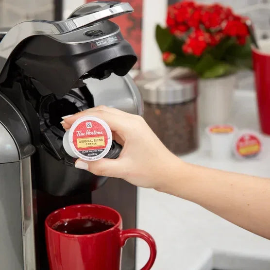 Hand inserting Tim Hortons Original Blend coffee pod into single-serve brewer with red mug nearby