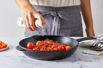 Hand spraying cooking oil on fresh cherry tomatoes in a cast iron skillet on kitchen counter