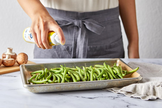 Person spraying cooking oil on fresh green beans in a baking tray, mushrooms on cutting board