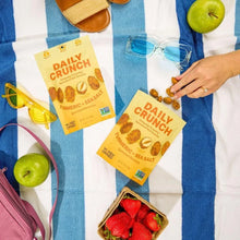 Daily Crunch turmeric sea salt sprouted almonds snack packs on striped picnic blanket with apples, strawberries, sunglasses, and sandals.