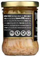 A glass jar of Tuna Inverse Pressed on a white background, showing the contents of shredded tuna in oil, a gold metal lid, and a black label with nutritional facts and certification logos.