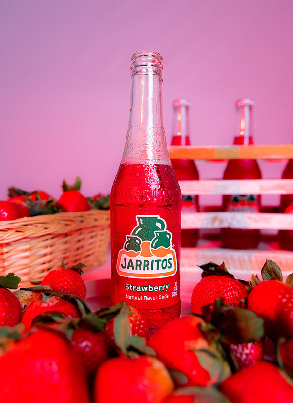 A bottle of Jarritos Strawberry natural flavor soda stands in the center, surrounded by fresh red strawberries and a wicker basket, with more bottles blurred in the background against a pink backdrop.