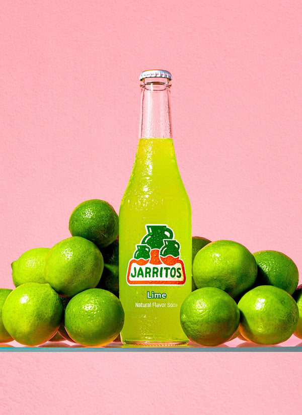 Jarritos lime soda bottle surrounded by limes on a pink background