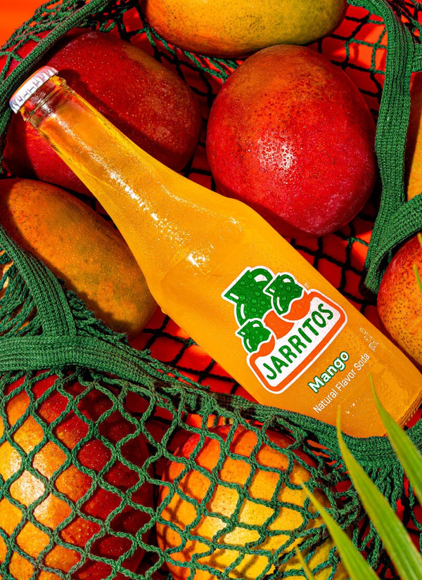 A bottle of Jarritos Mango natural fruit soda placed diagonally inside a green mesh bag filled with fresh red and yellow mangoes.