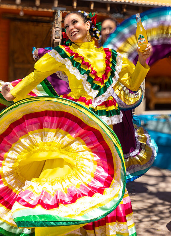A woman in a vibrant traditional Mexican-style dress with a large yellow, pink, green, and white skirt twirls outdoors while holding a bottle of beer. The dress features layered ruffles and bright, saturated colors.